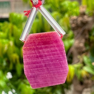 May include: A faceted, rectangular pink gemstone pendant with a red cord and clear glass accents. The pendant is a vibrant magenta color with visible striations. The background is blurred green foliage.