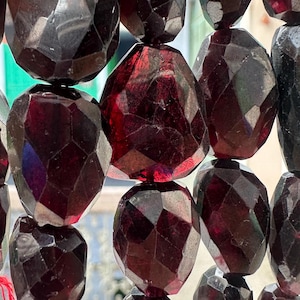 Peut inclure: Un collier de perles de grenat rouge foncé à facettes, présentant une forme irrégulière et organique. Les pierres sont enfilées sur un fil rouge fin, révélant une qualité sombre et translucide avec de multiples surfaces polies qui reflètent la lumière.