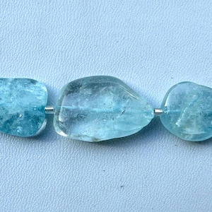 Könnte beinhalten: Eine Kette aus Aquamarin-Edelsteinperlen. Die hellblauen Perlen haben unregelmäßige Formen und eine polierte Oberfläche. Jede Perle ist auf einem dünnen, silberfarbenen Draht aufgefädelt.