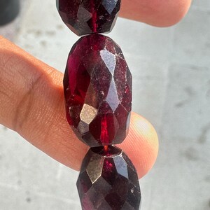 Puede incluir: Primer plano de una hebra de cuentas de piedras preciosas facetadas de color rojo oscuro. Las cuentas son de forma ovalada y tienen un acabado brillante.