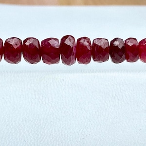 Könnte beinhalten: Eine Kette aus facettierten, tiefroten Rubinperlen. Die Perlen sind gleichmäßig groß und aufgereiht, wodurch eine durchgehende Farblinie entsteht. Die Edelsteine haben eine polierte, reflektierende Oberfläche, vor weißem Hintergrund.