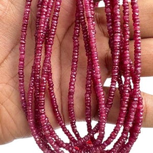 May include: Strands of small, faceted ruby beads in varying shades of red and pink. The beads are strung together, creating multiple strands. The gemstones are held in a hand, showcasing their color and texture. Ideal for jewelry making.