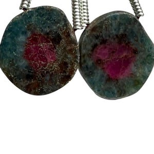 Könnte beinhalten: Zwei natürliche Edelsteinanhänger mit einer türkis-braunen Matrix, jeder mit einem leuchtenden magenta Zentrum. Die Anhänger sind an silberfarbenen, spiralförmigen Metallsträngen aufgehängt, ideal für die Schmuckherstellung.