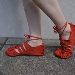 May include: A pair of red suede sandals with a lace-up design. The sandals have a flat sole and are tied around the ankles with matching red laces. The shoes are a vibrant shade of red.