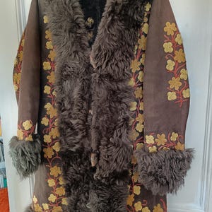 Op de afbeelding: Een bruine suède jas met een kraag, manchetten en bies van imitatiebont. De jas heeft geborduurde gele en rode bloempatronen langs de mouwen en de voorkant. De jas hangt aan een zwarte hanger.