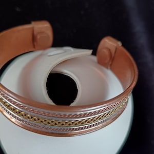 Puede incluir: Pulsera magnética de cobre con bandas plateadas y doradas. La pulsera abierta tiene dos extremos redondeados. La pulsera está sobre una superficie blanca sobre un fondo oscuro.