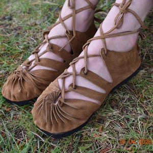 May include: Brown suede sandals with a lace-up design. The sandals have a flat sole and a gathered detail at the toe. The laces wrap around the ankle and up the leg. The sandals are on a grassy surface.