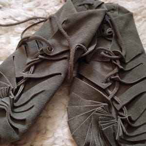 May include: A pair of olive green suede moccasins with intricate cut-out designs and laced closures. The shoes are displayed on a textured, off-white surface, showcasing their handcrafted details and earthy tones.