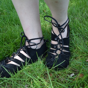 Puede incluir: Un par de sandalias negras de encaje con recortes. Las sandalias son de gamuza y tienen la punta cerrada. Las sandalias se atan al tobillo con cordones.