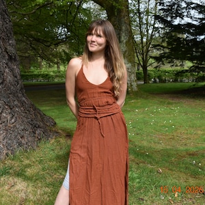 Puede incluir: Una mujer con un vestido largo color óxido con cuello halter y cintura ceñida. El vestido tiene una abertura lateral. La mujer está de pie en una zona de césped con árboles al fondo.