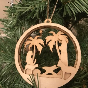 May include: A wooden Christmas ornament featuring a nativity scene with palm trees, a star, and a manger. The ornament is cut out and has a rustic brown color.