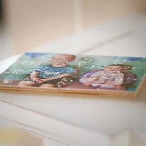 May include: A wooden jigsaw puzzle with a photo of two children. The older child is wearing a blue shirt with the word "BIG" on it. The younger child is wearing a white shirt with a floral pattern. The puzzle is assembled and sitting on a white surface.