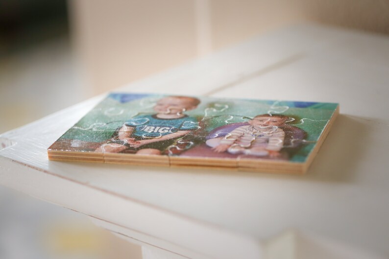 May include: A wooden jigsaw puzzle with a photo of two children. The older child is wearing a blue shirt with the word "BIG" on it. The younger child is wearing a white shirt with a floral pattern. The puzzle is assembled and sitting on a white surface.