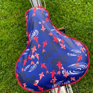 May include: A blue bicycle seat cover with a red border. The cover features a pattern of red and white fish swimming in blue water with white ripples.