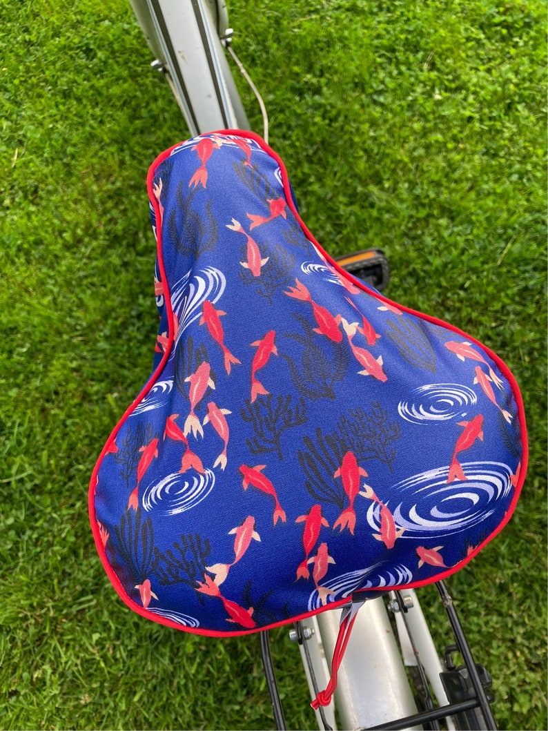 May include: A blue bicycle seat cover with a red border. The cover features a pattern of red and white fish swimming in blue water with white ripples.