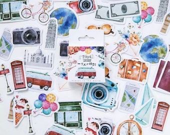 Op de afbeelding: Een set kleurrijke stickers met een reisthema met afbeeldingen van een vintage busje, een camera, een kompas, een koffer, een globe, een luchtballon, een telefooncel en een gebouw met een bord waarop "Metro" staat. De stickers zitten in een doorzichtige plastic doos met de tekst "Trend" en "Travel to the World" op de voorkant.
