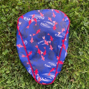 May include: A blue bicycle seat cover with a red border. The cover features a pattern of red fish swimming in blue water with white ripples.