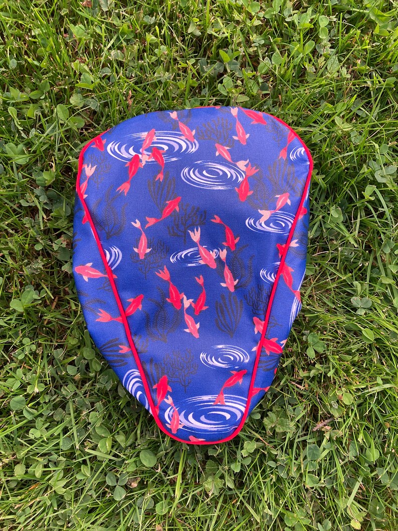 May include: A blue bicycle seat cover with a red border. The cover features a pattern of red fish swimming in blue water with white ripples.
