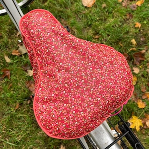 Op de afbeelding: Een rode fietszadelhoes met een bloemenpatroon. De hoes heeft een roze rand en staat tegen een achtergrond van groen gras en gevallen bladeren. De zitting is bevestigd aan een zilveren fiets.
