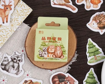Op de afbeelding: Een set kleurrijke stickers met verschillende bosdieren, waaronder een wasbeer, een uil, een hert, een egel en een rode panda. De stickers bevinden zich op een houten oppervlak en zijn verpakt in een klein doosje met de tekst "Jungle Family" in het Chinees en Engels.