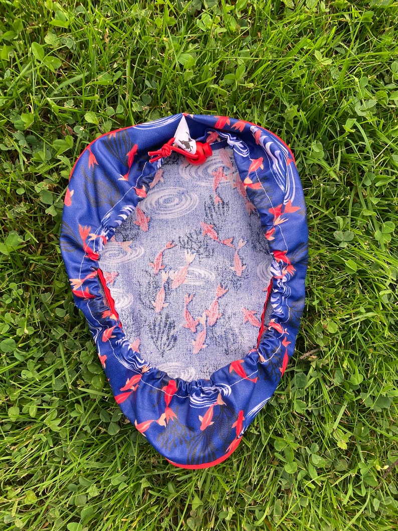 May include: A blue and red fabric cover with a white drawstring. The fabric has a pattern of red fish swimming in blue water with white waves.