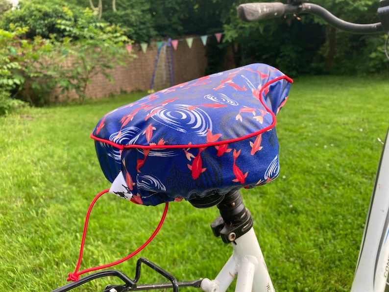 May include: A blue bicycle seat cover with a red trim and a pattern of red fish swimming in blue water. The cover has a red drawstring.
