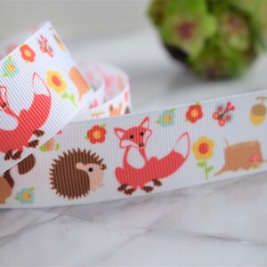 May include: A white grosgrain ribbon featuring a repeating pattern of orange foxes, brown hedgehogs, and light brown squirrels interspersed with colourful flowers, green leaves, and small brown acorns on a textured fabric surface.