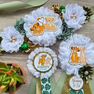 May include: A set of baby shower decorations featuring The Lion King theme. The set includes a sash with white fabric flowers, green and gold accents, and two rosettes with the text "Daddy To Be" and "Mommy To Be".