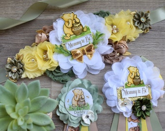 May include: A maternity sash and two pins featuring Winnie the Pooh illustrations. The sash has white and yellow fabric flowers, a gold bow, and a sign reading Mommy to Be. The pins feature Winnie the Pooh with a honey pot and text reading Mommy to Be or Daddy to Be.