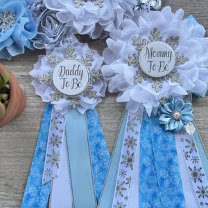 以下が含まれることがあります： 「Daddy To Be」と「Mommy To Be」の文字が入った、白と水色のベビーシャワーリボン。リボンには雪の結晶と花の模様があります。青い花と多肉植物も見えます。