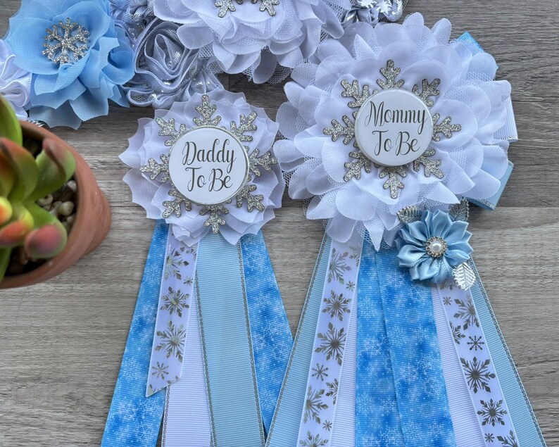 以下が含まれることがあります： 「Daddy To Be」と「Mommy To Be」の文字が入った、白と水色のベビーシャワーリボン。リボンには雪の結晶と花の模様があります。青い花と多肉植物も見えます。