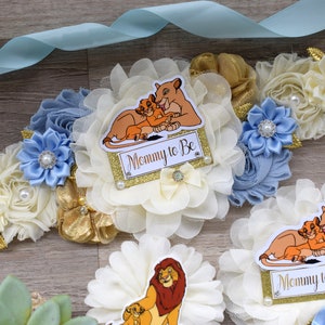 Pu&ograve; includere: Nastro azzurro con decorazioni floreali color crema, oro e blu. Il design include un cartello "Mommy to Be" con una grafica del Re Leone, perle e accenti decorativi. Tema baby shower.
