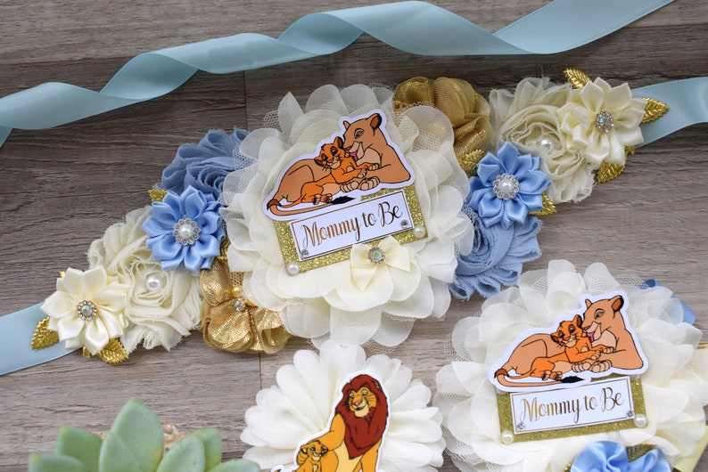 Pu&ograve; includere: Nastro azzurro con decorazioni floreali color crema, oro e blu. Il design include un cartello "Mommy to Be" con una grafica del Re Leone, perle e accenti decorativi. Tema baby shower.