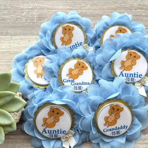 Pu&ograve; includere: Spille per baby shower a forma di fiore blu chiaro, con un'immagine di cucciolo di leone dei cartoni animati e bordi dorati. Ogni spilla ha titoli come "Zia -TO BE-" e "Nonno -TO BE-". Alcune hanno un fiocco bianco.