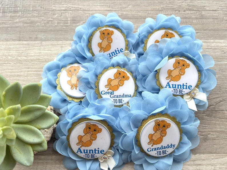 Pu&ograve; includere: Spille per baby shower a forma di fiore blu chiaro, con un'immagine di cucciolo di leone dei cartoni animati e bordi dorati. Ogni spilla ha titoli come "Zia -TO BE-" e "Nonno -TO BE-". Alcune hanno un fiocco bianco.