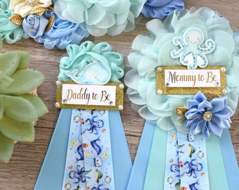 May include: Two blue and green floral sashes with a gold glitter background. One sash says "Daddy to Be" and the other says "Mommy to Be". The sashes are decorated with a whale and an octopus.