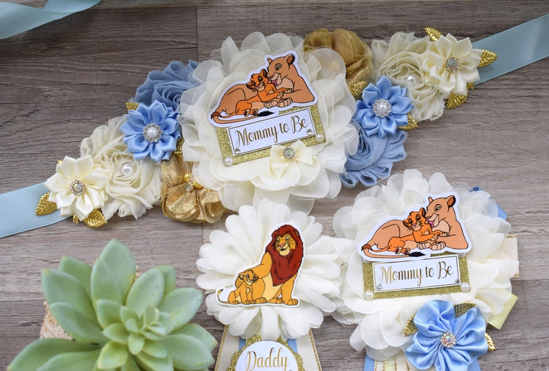 Pu&ograve; includere: Una fascia floreale blu e bianca con dettagli dorati e un distintivo "Mommy to Be" con una leonessa e il suo cucciolo. Sono visibili anche altri distintivi a tema Re Leone, perfetti per un baby shower.