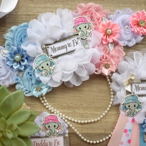 May include: Two baby shower sashes with blue and pink elephant designs. The sashes are decorated with white, blue, and pink flowers and have the text "Mommy to Be" on them.