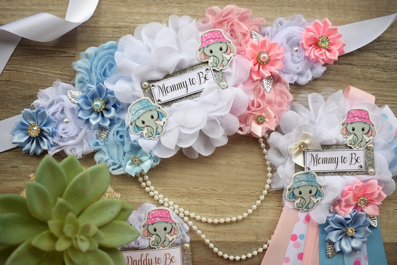 May include: Two baby shower sashes with blue and pink elephant designs. The sashes are decorated with white, blue, and pink flowers and have the text "Mommy to Be" on them.