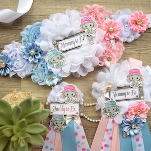 May include: A set of three baby shower sashes with blue and pink ribbons, white flowers, and elephant appliques. The sashes have the text "Mommy to Be" and "Daddy to Be" printed on them.