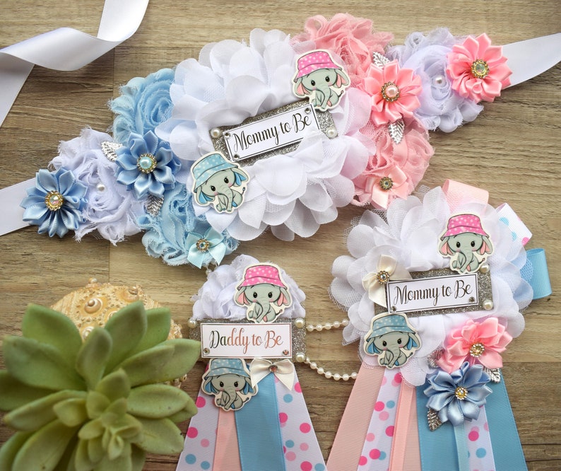 May include: A set of three baby shower sashes with blue and pink ribbons, white flowers, and elephant appliques. The sashes have the text "Mommy to Be" and "Daddy to Be" printed on them.