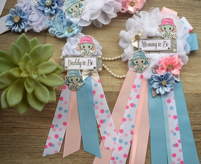 May include: Two blue and pink sashes with white polka dots and a white, blue, and pink flower embellishment. Each sash has a small elephant with a hat and a sign that says "Mommy to Be" and "Daddy to Be".