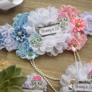 May include: A white, blue, and pink fabric sash with a silver glitter 'Mommy to Be' sign. The sash is decorated with flowers, bows, and elephant appliques.