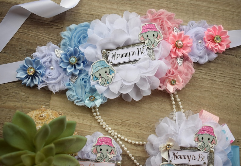 May include: A white, blue, and pink fabric sash with a silver glitter 'Mommy to Be' sign. The sash is decorated with flowers, bows, and elephant appliques.