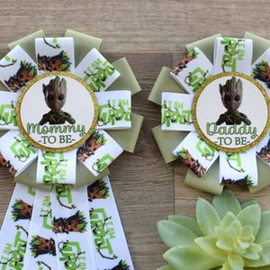 May include: Two decorative baby shower rosettes. One says "Mommy -TO BE-" and the other says "Daddy -TO BE-". Both have a gold-rimmed center with a Groot character image. The ribbons are white and green.