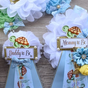 May include: Two blue and white baby shower sashes with a turtle design. One sash says "Mommy to Be" and the other says "Daddy to Be".