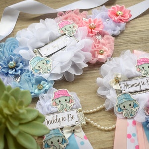 May include: Three baby shower sashes with blue and pink decorations. The sashes have the text "Mommy to Be", "Daddy to Be", and "Mommy to Be" on them. Each sash has a small elephant with a hat on it.