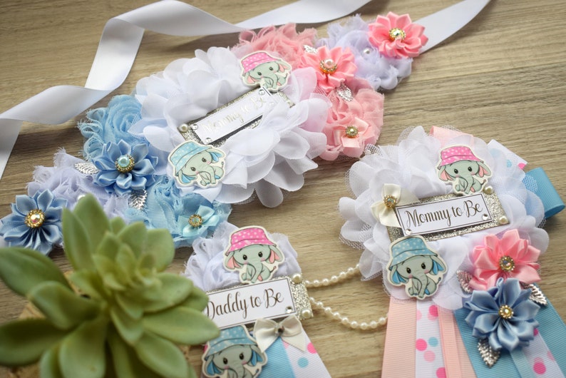 May include: Three baby shower sashes with blue and pink decorations. The sashes have the text "Mommy to Be", "Daddy to Be", and "Mommy to Be" on them. Each sash has a small elephant with a hat on it.