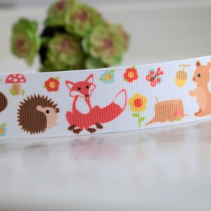 May include: A white grosgrain ribbon featuring a repeating pattern of woodland animals including brown squirrels, brown hedgehogs, and orange foxes, interspersed with colourful red mushrooms, yellow flowers, and brown acorns.