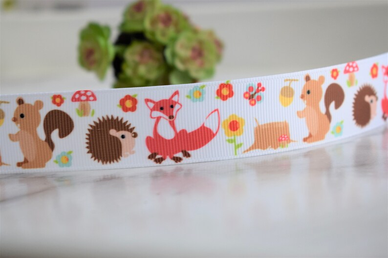 May include: A white grosgrain ribbon featuring a repeating pattern of woodland animals including brown squirrels, brown hedgehogs, and orange foxes, interspersed with colourful red mushrooms, yellow flowers, and brown acorns.